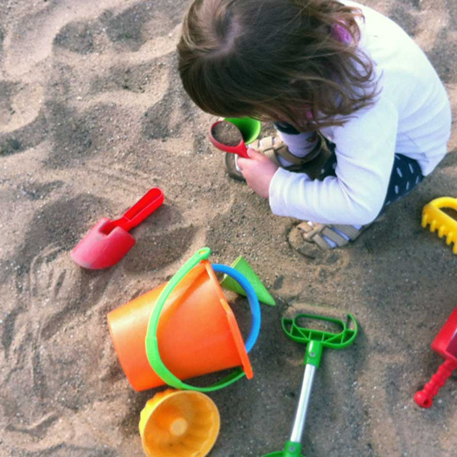 Ein Kind spielt konzentriert im Sand mit buntem Sandspielzeug, darunter ein orangener Eimer, rote und grüne Schaufeln sowie eine gelbe Sandschale. Das Kind trägt eine weiße Oberbekleidung, dunkle Hosen mit Punkten und Sandalen, während es mit einer kleinen Schaufel Sand untersucht. Die Szene vermittelt spielerische Kreativität und Entdeckerfreude.