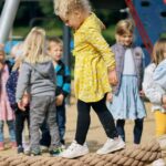 Eine Gruppe von Kindern spielt auf einem Spielplatz. Im Vordergrund balanciert ein Mädchen in einem gelben Kleid auf einem dicken Seil, während im Hintergrund andere Kinder warten oder auf Spielgeräten klettern. Die Szene zeigt Freude und spielerisches Miteinander in einer entspannten Umgebung.