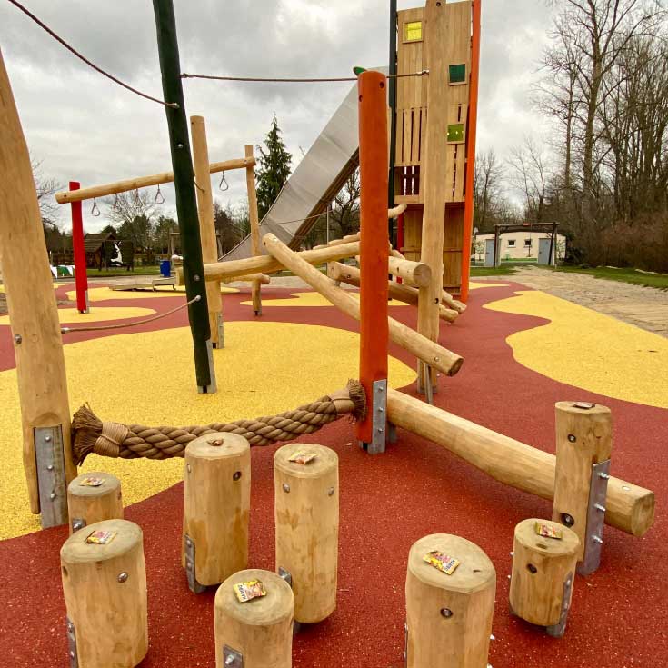 Ein moderner Spielplatz mit natürlichen Holzelementen. Im Vordergrund stehen Holzpfosten als Balancierpfad, verbunden mit einem dicken Seil. Im Hintergrund sind ein großer Kletterturm mit Rutsche und weitere Kletterstrukturen zu sehen. Der Boden ist mit einer rot-gelben Fallschutzfläche gestaltet, während kahle Bäume und eine bewölkte Himmelsszenerie die Umgebung bestimmen.